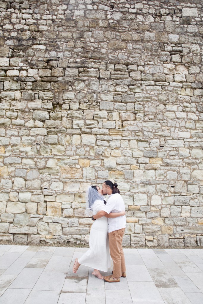 A bride in a white dress and veil kisses her groom, who is dressed casually in a white shirt and brown trousers, in front of a rustic stone wall after their registry office wedding in Southampton.