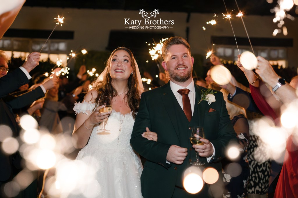 Bride and groom smiling during sparkler exit at wedding in the New Forest, Burley Manor, photographed by Katy Brothers Wedding Photography, capturing joyful, natural moments.